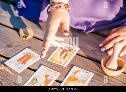 Die Hand der Frau mit violetten Nägeln zeigt auf vier Tarot-Karten, die auf einer Holzfläche neben Stacheletts und Lavendel verteilt sind. Minsk, Weißrussland - 07.28.2021 Stockfoto