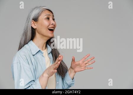 Überraschte weißhaarige Frau, die gestikulierte und isoliert über die graue Wand schaute Stockfoto