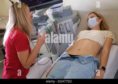 Frau in schützender medizinischer Maske, die mit Ultraschalldiagnostik auf der Couch vor dem Arzt liegt Stockfoto