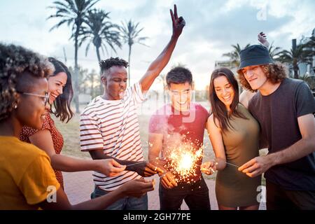 Eine Gruppe von Freunden zünden bei Sonnenuntergang einige Wunderkerzen an. Junge Multiethniker feiern gemeinsam. Konzept von Sommer, Integration, Glück, Freizeit Stockfoto