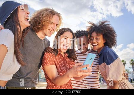 Eine Gruppe junger Menschen, die Spaß daran haben, etwas auf ihrem Handy zu sehen. Die Jungen umarmen und lachen miteinander. Konzept der multiethnischen, Freundschaft Stockfoto