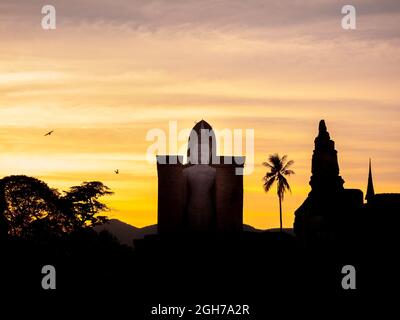 Atemberaubende Silhouette des Wat Mahathat Tempels im Bezirk des Sukhothai Historical Park mit einer großen buddha-Statue auf einem goldenen Himmel bei Sonnenuntergang Stockfoto