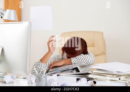 Erschöpfte junge Geschäftsfrau, die mit dem Gesicht auf dem Stapel von Dokumenten am Tisch sitzt und weiße Flagge zeigt Stockfoto