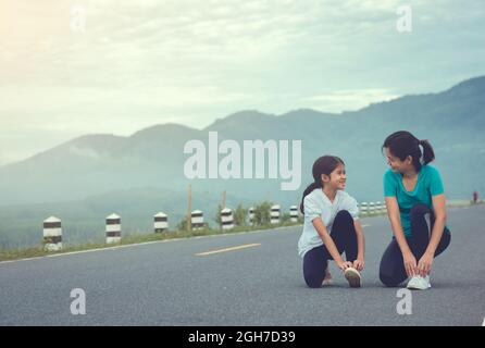 Mutter und Kind knien und binden Schnürsenkel. Wir machen uns bereit zum Joggen im Freien während des Sonnenaufgangs auf der Staudamm-Straße zu trainieren. Stockfoto