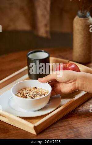 Nahaufnahme einer Frau, die Frühstück zubereitet und eine Schüssel mit Müsli, Apfel und einer Tasse Kaffee auf das Tablett stellt Stockfoto