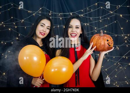 Junge kaukasische Frau, die Kürbis in ein Gesicht gehauen hält. Ihre Freundin hält Luftballons in der Hand. Beide genießen halloween-Party mit blinkenden Lichtern geschmückt. Stockfoto