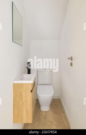 Schmale weiße Toilette mit kleinem Waschbecken, Spiegel und Holzmöbeln. Stockfoto