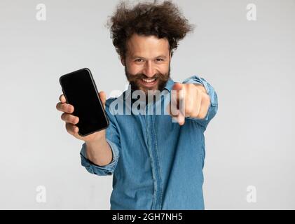 Hübscher Mann mit Schnurrbart, der die Arme nach oben hält, mit dem Finger zeigt und im Studio auf grauem Hintergrund einen leeren schwarzen mobilen Bildschirm zeigt Stockfoto