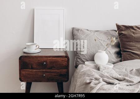 Vertikales Modell mit weißem Bilderrahmen. Nachttisch aus Holz im Retro-Stil. Moderne weiße Keramikvase mit trockenem Lagurus ovatus Gras und einer Tasse Kaffee. Beige Stockfoto