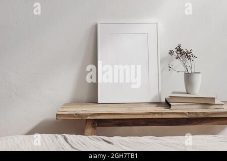 Vertikales Modell mit weißem Bilderrahmen. Vintage Holzbank, Tisch. Becher mit trockenem Gras auf Bücherhaufen. Weißer Wandhintergrund. Skandinavische Innenausstattung Stockfoto