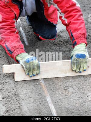 Ein kniender Arbeiter in roten Overalls ebnet das sandige Fundament mit einer Holzebene für zukünftige Gehwegverkleidungen. Speicherplatz kopieren. Vertikales Bild. Stockfoto