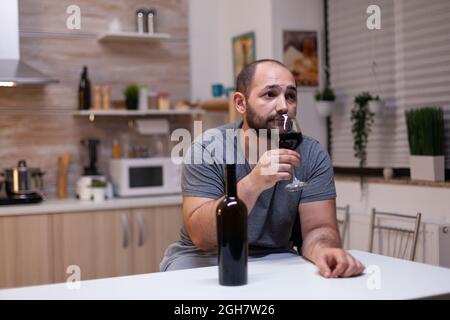 Kaukasischer Mann hält ein Glas Wein in der Küche zu Hause. Depressive Person, die Alkohol trinkt, Alkohol trinkt, alkoholisches Getränk allein, während sie sich mit einer Flasche Alkohol berauscht fühlt Stockfoto