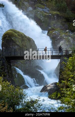 Touristen überqueren eine Brücke vor einem tosenden Wasserfall am Briskdal Gletscher, Jostedal Gletscher Nationalpark, Norwegen, Europa Stockfoto