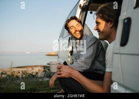 Lächelndes, schwules Paar, das am Seeufer sitzt und Kaffee getrunken hat Stockfoto