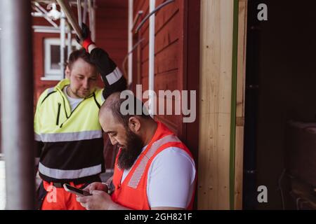 Männlicher Bauarbeiter, der auf der Baustelle einen bärtigen Kollegen mit einem Smartphone ansieht Stockfoto