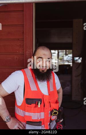 Lächelnder bärtiger Bauarbeiter, der auf der Baustelle mit der Hand auf der Hüfte stand Stockfoto