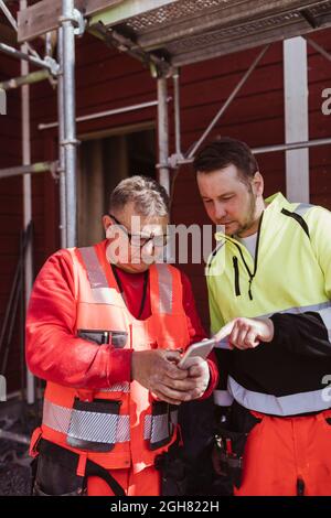 Reifer männlicher Bauarbeiter, der mit einem Kollegen auf der Baustelle über ein Smartphone diskutiert Stockfoto