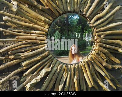 Ein Mädchen, das im Park spazieren geht, spiegelt sich in einem runden Spiegel wider, der mit Holzstücken geschmückt ist. Sonnenförmiger Parkspiegel Stockfoto