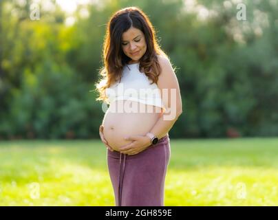 Porträt einer Schwangeren, die ihren Bauch berührt, während sie im Freien nach unten schaut Stockfoto