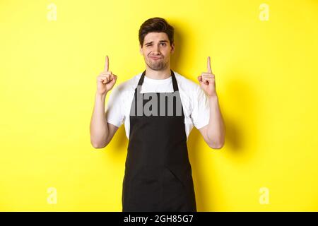 Unzufriedener und skeptischer Kellner mit schwarzer Schürze, der die Finger nach oben zeigt, drückt Abneigung und Gleichgültigkeit aus und steht auf gelbem Hintergrund Stockfoto