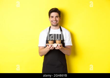 Ein hübscher Barista, der Kaffee zum Mitnehmen serviert und lächelt und Ordnung bringt. Er steht in einer schwarzen Schürze vor gelbem Hintergrund Stockfoto