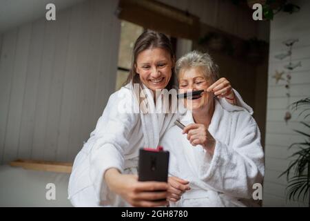 Glückliche ältere Mutter im Bademantel mit erwachsenen Tochter, die zuhause Selfie macht, Selfcare-Konzept. Stockfoto