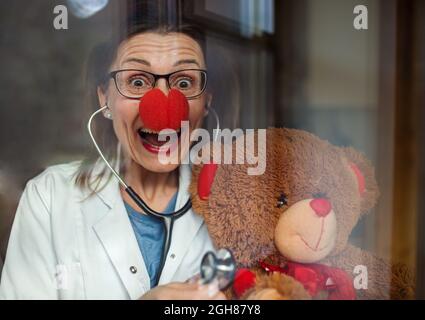 Porträt einer lächelnden Ärztin mit roter Clownnase, die einen Teddybären hält und die Kamera anschaut. Stockfoto