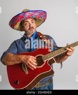 Emotionaler mexikanischer Erwachsener im nationalen Sombrero-Kopfschmuck spielt Gitarre auf grauem Hintergrund Stockfoto