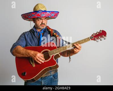 Emotionaler mexikanischer Erwachsener im nationalen Sombrero-Kopfschmuck spielt Gitarre auf grauem Hintergrund Stockfoto