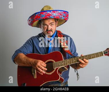 Emotionaler mexikanischer Erwachsener im nationalen Sombrero-Kopfschmuck spielt Gitarre auf grauem Hintergrund Stockfoto