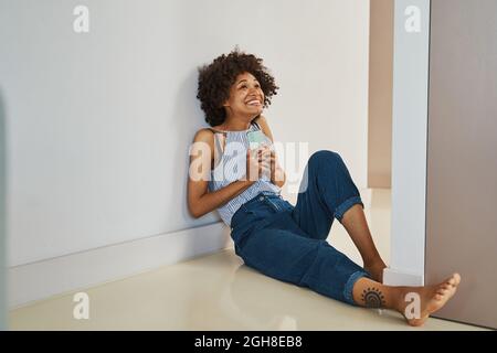 Fröhliche Dame mit dem Smartphone an der Wand sitzend und nach oben schauend Stockfoto