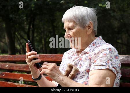 Ältere Frau, die mit dem Smartphone auf einer Bank im Park sitzt. Online-Kommunikation, sms, Leben im Ruhestand Stockfoto