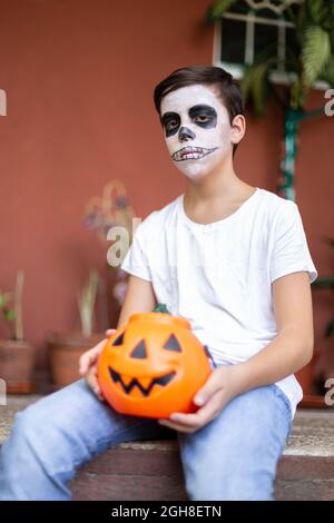 Porträt eines Mannes mit Gesicht als Schädel, der im Eingang seines Hauses sitzt. Er ist sehr ernst und hält einen Halloween Kürbis. Stockfoto