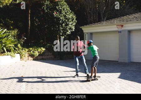 afroamerikanischer Vater lächelt und hilft Sohn balanciert auf Skateboard im Garten Stockfoto