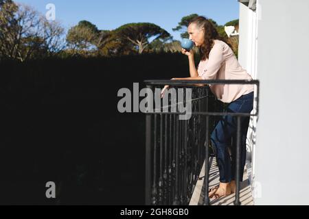 Glückliche ältere kaukasische Frau, die Kaffee trinkt und auf dem Balkon steht Stockfoto