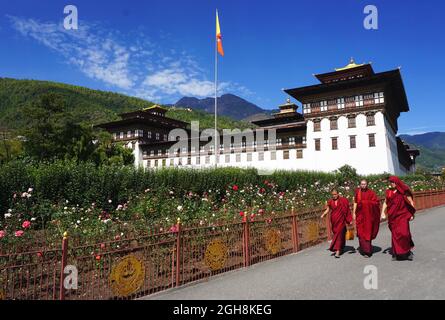 Drei buddhistische Mönche mit roten Roben gehen an wunderschönen Blumengärten vor dem Tashichho Dzong vorbei, dem offiziellen Sitz der Regierung Bhutans seit 1952. Stockfoto