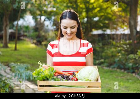 Lächelnde kaukasische Frau, die im Garten steht und eine Schachtel mit frischem Bio-Gemüse hält Stockfoto