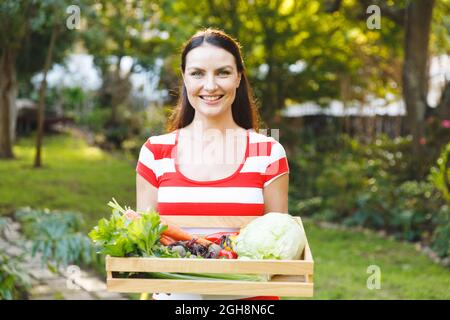 Porträt einer lächelnden kaukasischen Frau, die im Garten steht und eine Schachtel mit frischem Bio-Gemüse hält Stockfoto