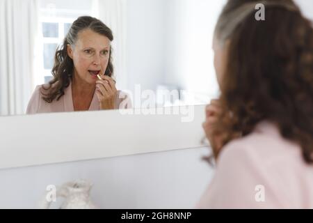 Glückliche ältere kaukasische Frau im Badezimmer, in Spiegel schauend, Lippenstift aufklebend Stockfoto