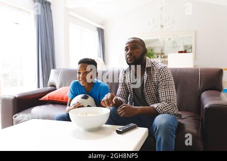 Glücklicher afroamerikanischer Vater und Sohn sitzen auf dem Sofa, schauen sich das Spiel im fernsehen an und sprechen Stockfoto