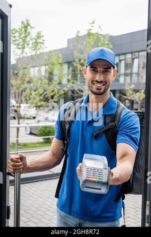 Fröhlicher muslimischer Kurier, der unscharfes Zahlungsterminal in der Nähe hält Stockfoto