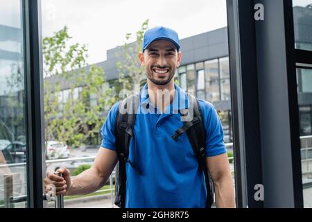 Lächelnder muslimischer Kurier mit Rucksack, der neben der Tür des Gebäudes steht Stockfoto