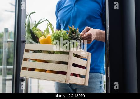 Beschnittene Ansicht des Lieferers, der eine Schachtel mit frischem Gemüse in der Nähe der Tür hält Stockfoto