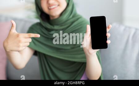 Nicht erkennbares muslimisches Teenager-Mädchen in Hijab, das zu Hause auf ein Mobiltelefon mit leerem Bildschirm zeigt, ein Modell für Design Stockfoto