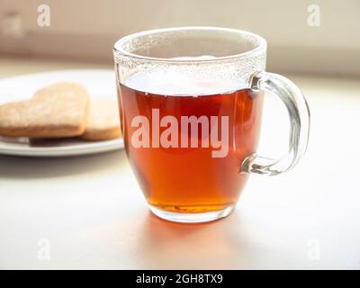 Komposition mit Tasse schwarzem Tee und Teller, Keks auf Fensterbank. Stillleben mit Becher und Getränk vor dem Fenster Stockfoto