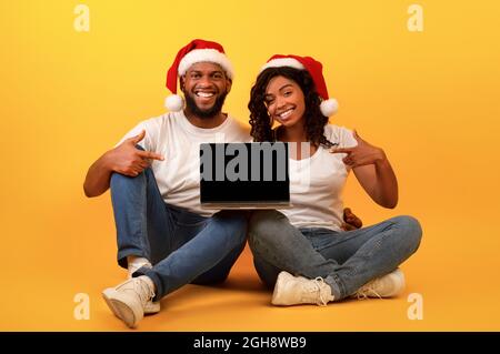 Aufgeregtes afroamerikanisches Paar in Weihnachtsmützen, das auf einen Laptop mit leerem Bildschirm zeigt und auf gelbem Hintergrund sitzt Stockfoto