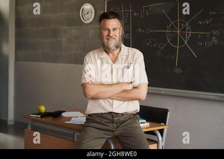 Der bärtige Lehrer mittleren Alters schaute mit gekreuzten Armen im Klassenzimmer auf die Kamera. Stockfoto