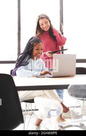 Vertikales Foto. Im Büro: Zwei Frauen arbeiten zusammen. Sie diskutieren Arbeitsprobleme freundlich, während sie auf den Computerbildschirm schauen. Stockfoto