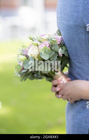 Nahaufnahme der Hand einer Braut mit einem Bouquet aus weißen und rosa Rosen, die die blaue Weste des Bräutigams auf einem Wiesenhintergrund umarmen. Hochwertige Fotos Stockfoto