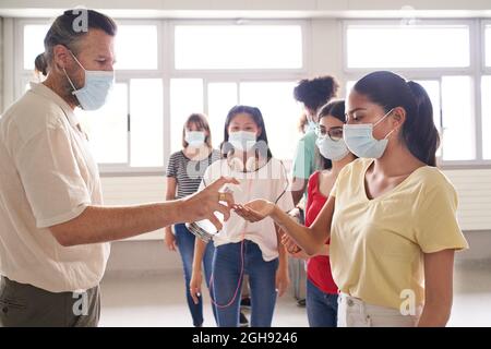 Eine Gruppe von Schülern in Gesichtsmasken wartet in der Schlange, um in den Klassenzimmer zu kommen. Der Lehrer wendet hydroalkoholisches Gel für die Virenschutzmaßnahmen an. Zurück zur Schule. Stockfoto